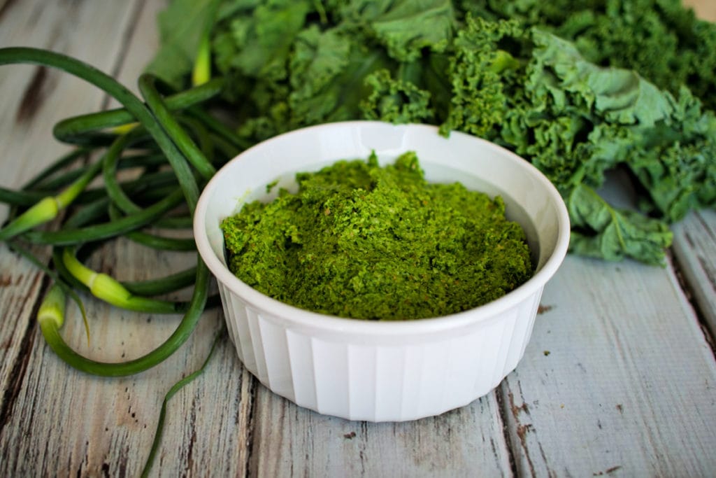 bowl of pesto in front of garlic scapes and kale