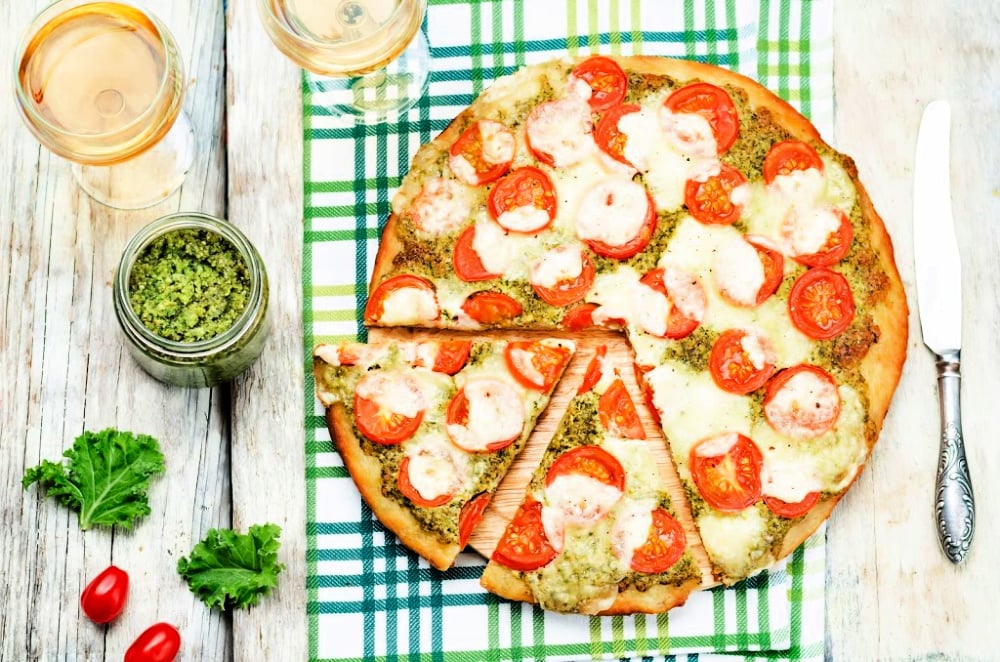 top view of a pesto pizza with mozzarella next to a jar of pesto, kale and wine glasses
