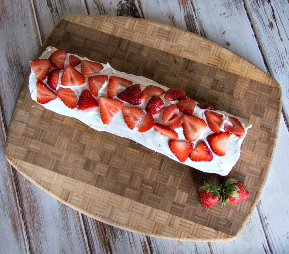 A strawberry roll cake with whipped cream and strawberries on a wooden cutting board.
