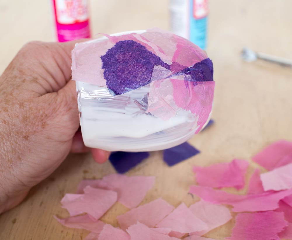 A person is placing tissue paper inside a vase to create stained glass votive candle holders.