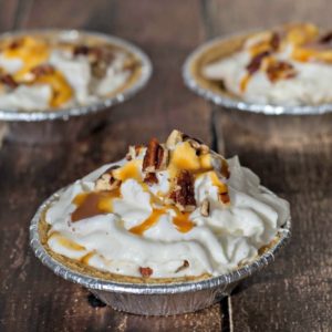 Three mini caramel apple pies with whipped cream and pecans on a wooden table.