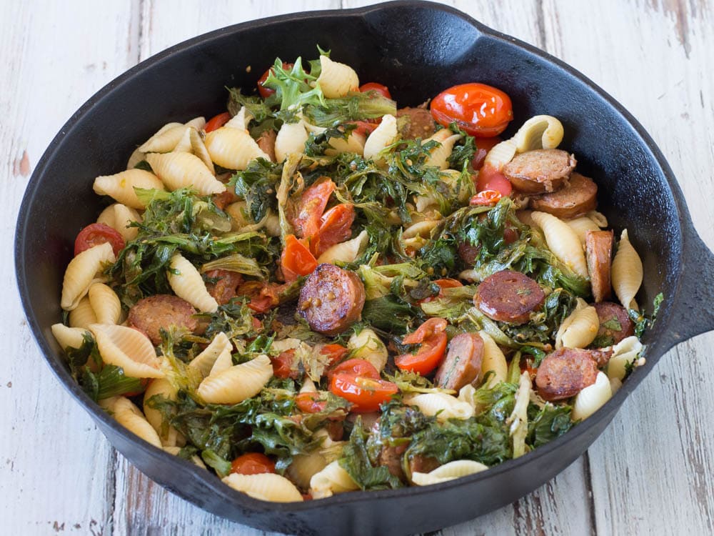 A skillet filled with pasta, sausage, arugula and greens.