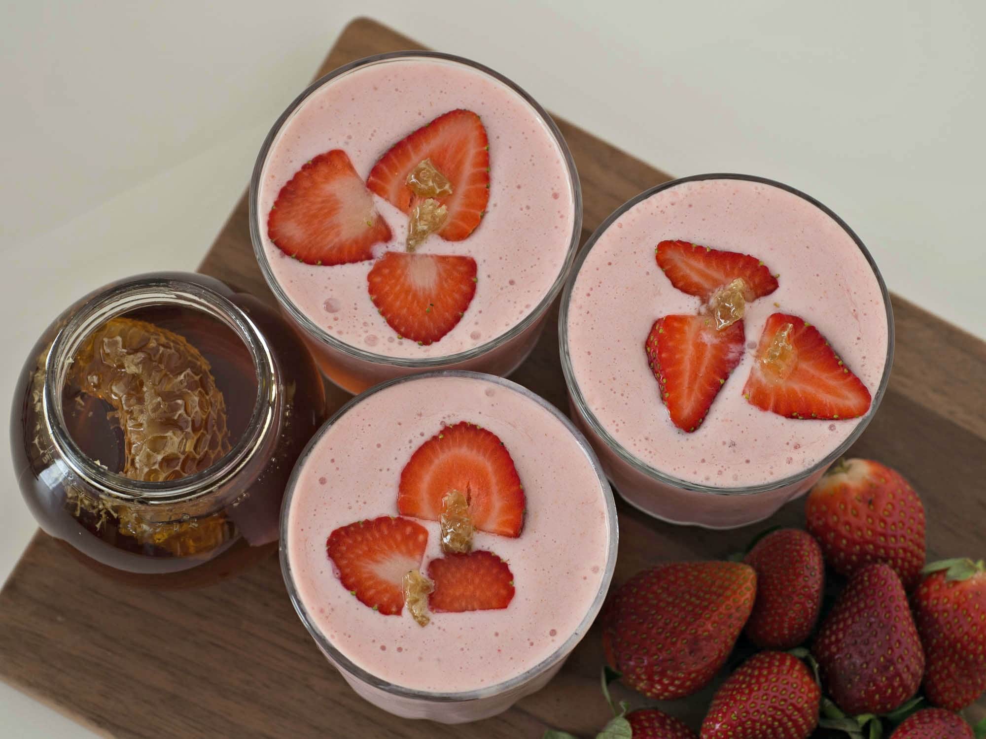 Three glasses of strawberry smoothie with honey on a cutting board.