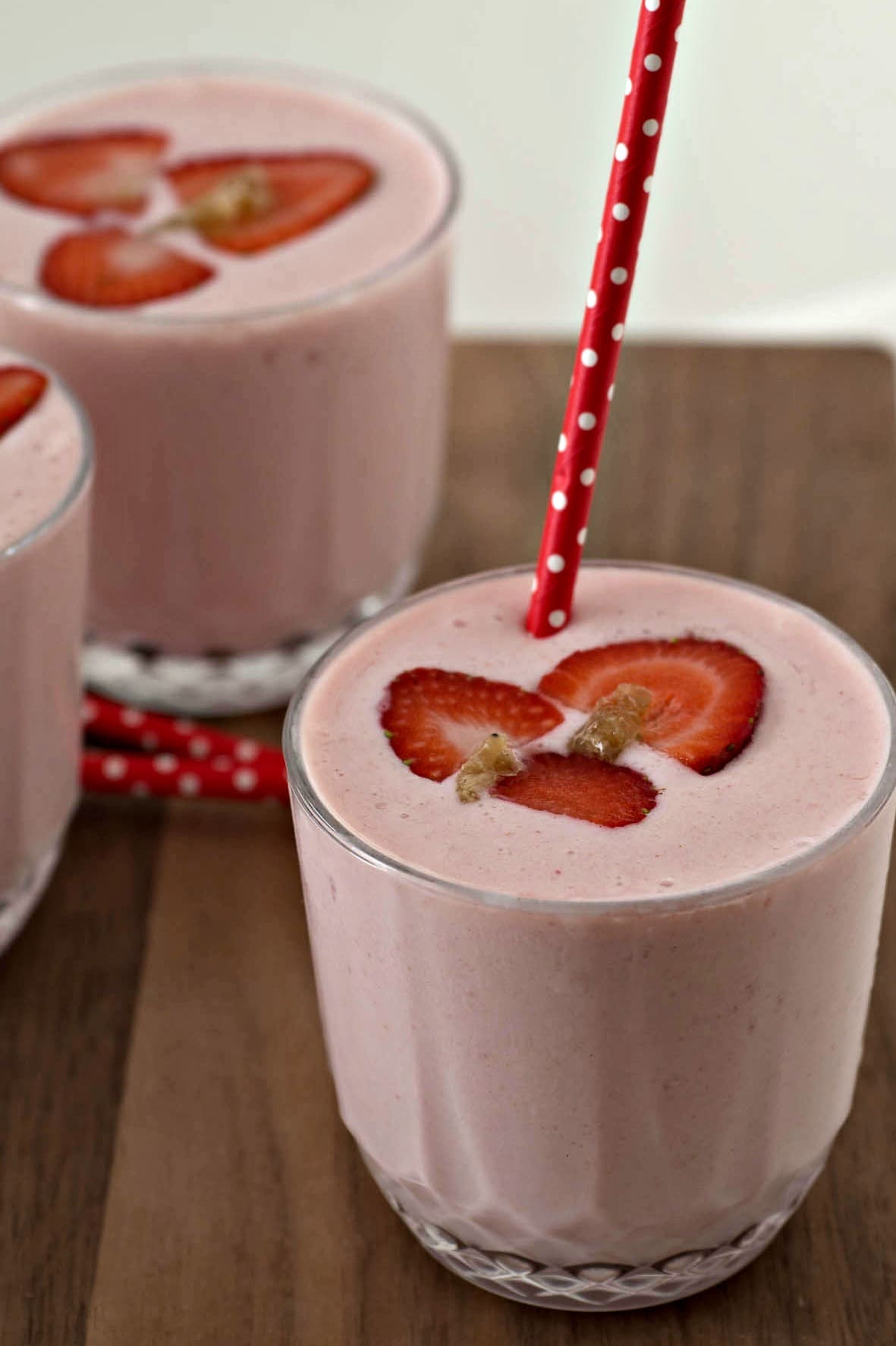 Three glasses of strawberry smoothie with straws on a cutting board.