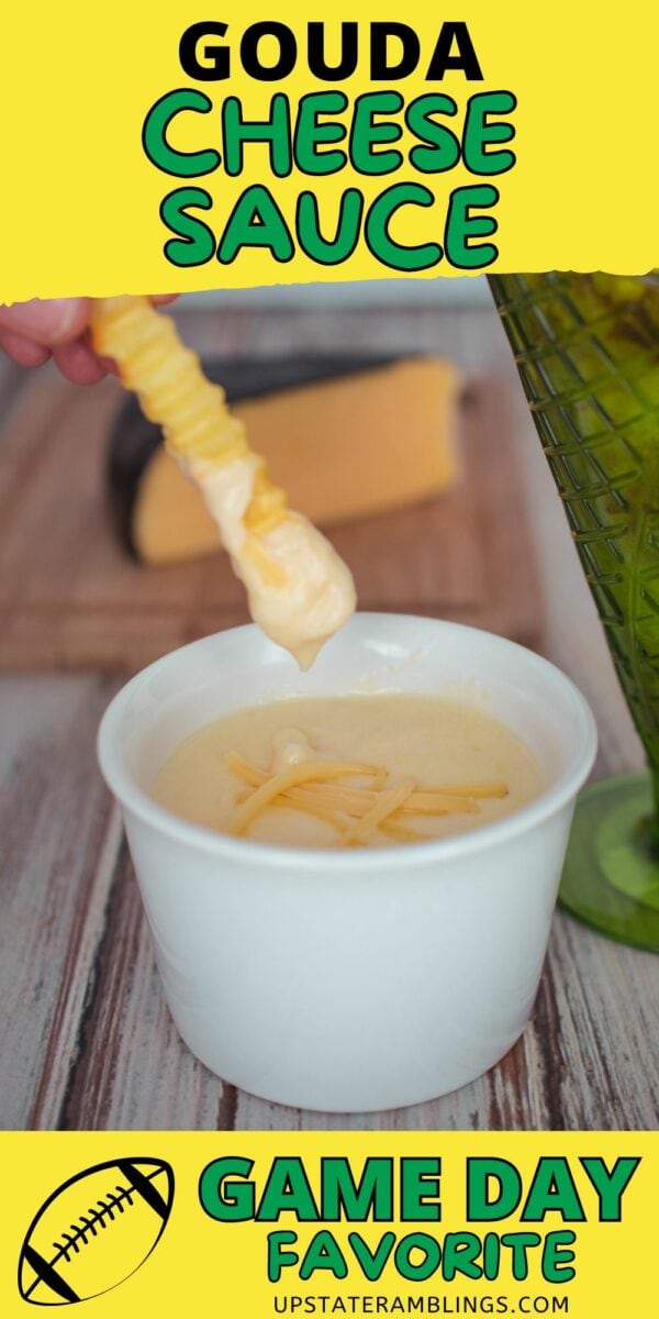 A crinkle-cut fry dipped in gouda cheese sauce is held above a white cup filled with the sauce. A wedge of gouda cheese and part of a green glass are visible in the background.