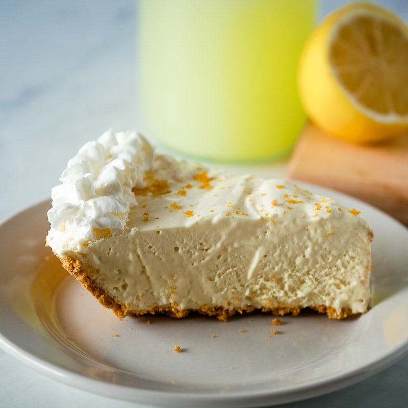 Slice of lemonade pie on a plate with lemonade in the background.