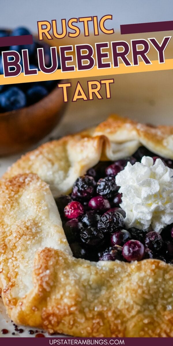 A rustic blueberry galette with a golden, flaky crust and a dollop of whipped cream on top. A bowl of fresh blueberries is visible in the background.