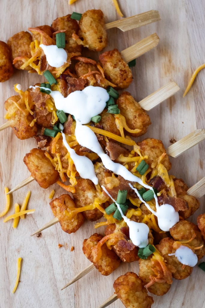tater tot skewers on a cutting board - topped with cheese, bacon, green onions and drizzled with ranch dressing