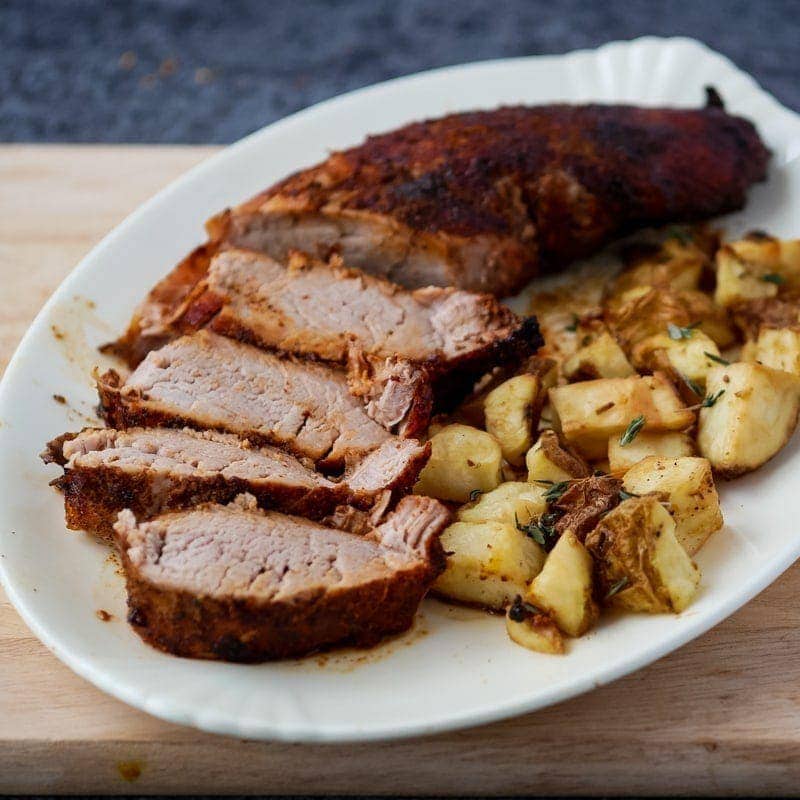 Air fryer pork tenderloin on a plate with potatoes on a cutting board.