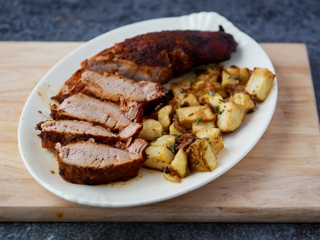Air fryer pork tenderloin on a plate with potatoes on a cutting board.