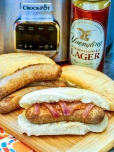 beer brats on a cutting board with beer and slow cooker in the background