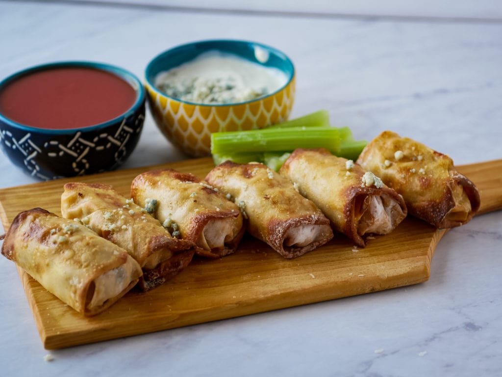 Buffalo chicken egg rolls on a wooden cutting board with sauce in the back