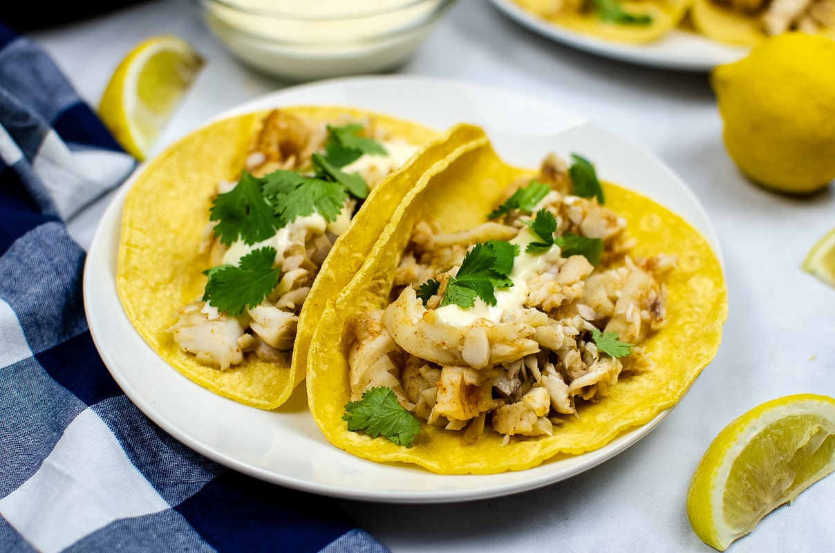 Fish tacos on a plate in tortillas with coriander and lemon wedges.
