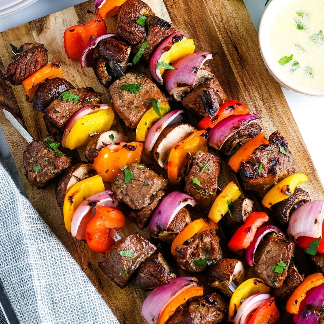 Steak kabobs with peppers and onions on a cutting board with sauce and plates in the background.