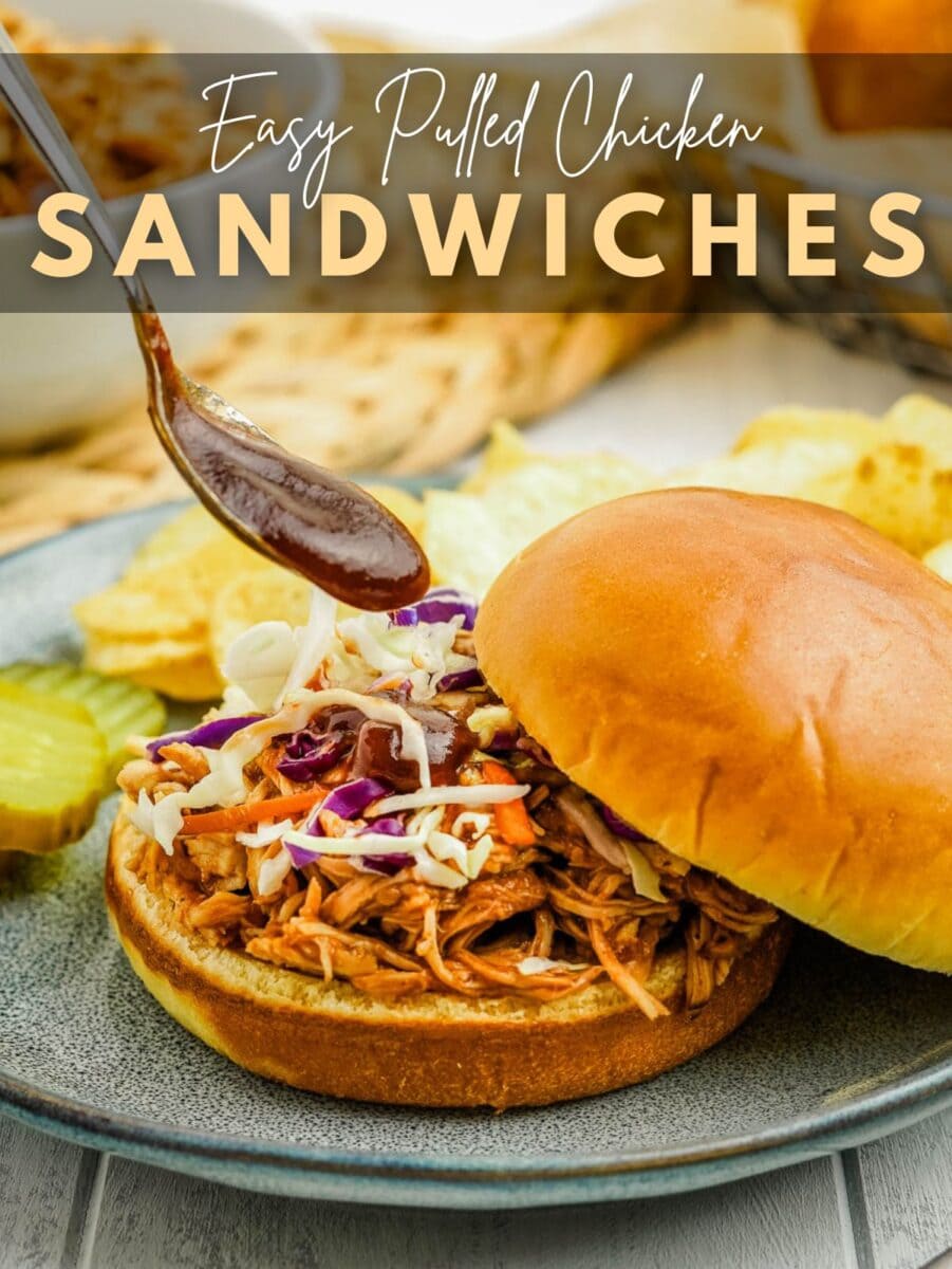 Slow cooker pulled chicken sandwich with coleslaw on a bun, served with pickle slices and potato chips, being topped with barbecue sauce.
