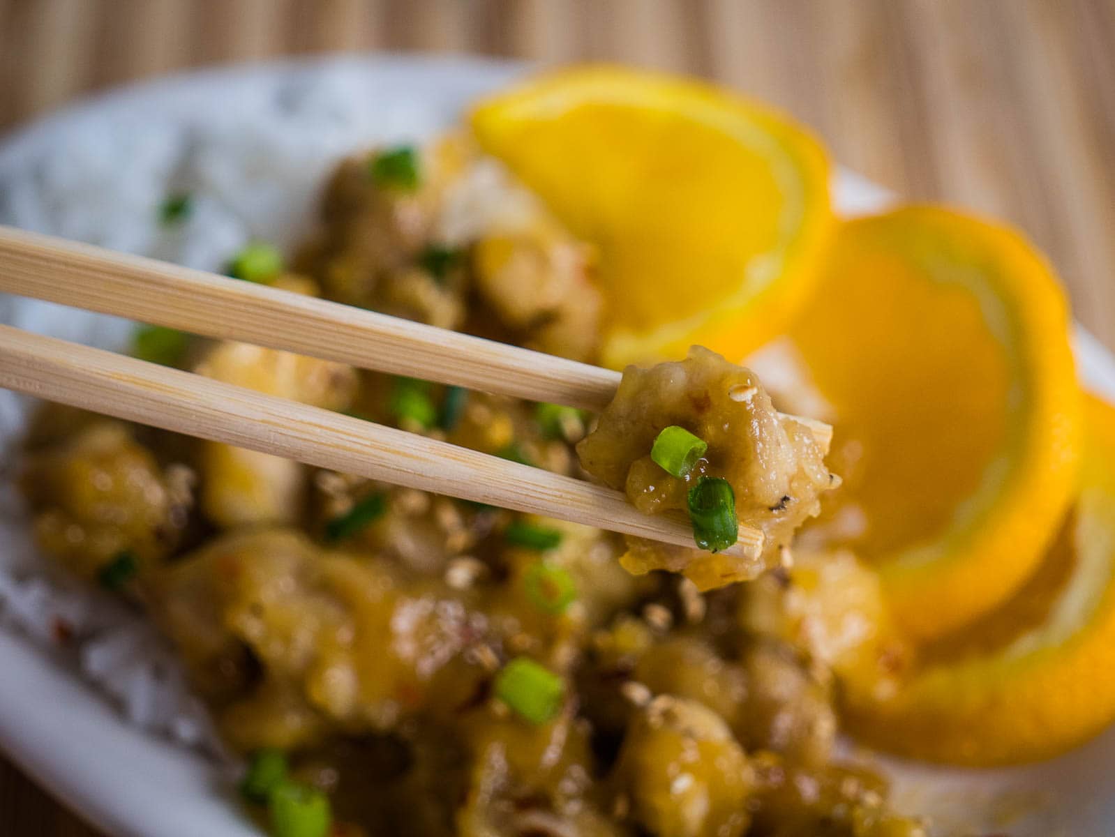 Orange chicken in chopsticks with chicken, rice and orange slices in the background.