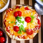 A freshly baked pizza topped with melted cheese, sliced red bell peppers, cherry tomatoes, green onions, and a dollop of Taco Bell's Mexican Pizza sauce, served on a wooden board.