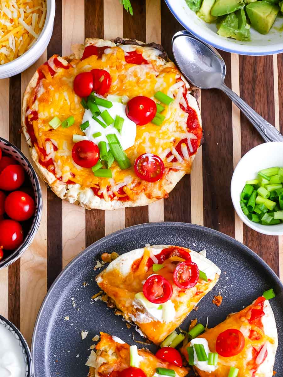 Taco Bell Mexican pizza topped with cheese, tomatoes, and green onions, served with side dishes of avocado, sour cream, and whole cherry tomatoes.