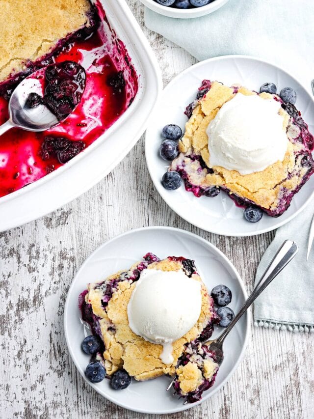 Two plates with blueberry cobbler topped with vanilla ice cream and fresh blueberries, next to a baking dish of cobbler on a white wooden surface.