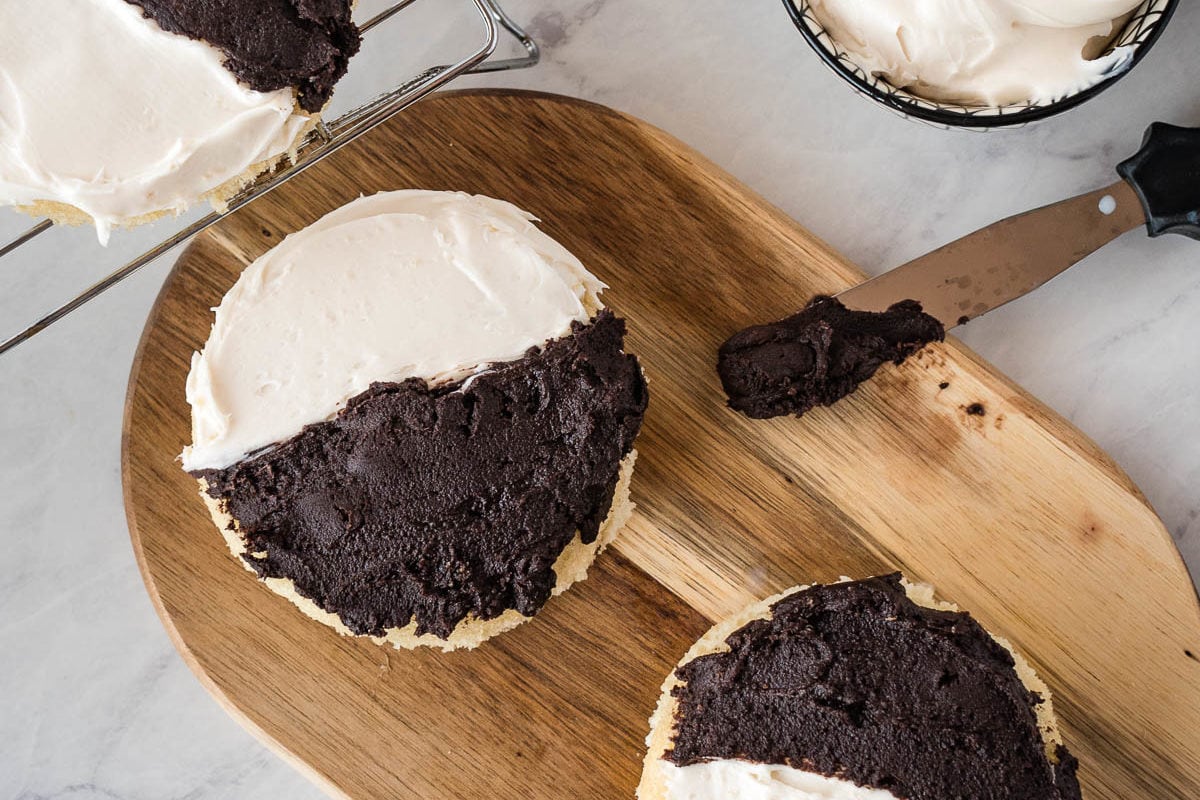 Half moon cookies on a wooden cutting board with ice with half vanilla and half chocolate frosting.