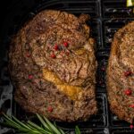 Two seasoned steaks with red peppercorns cooking in an air fryer, accompanied by fresh rosemary sprigs.