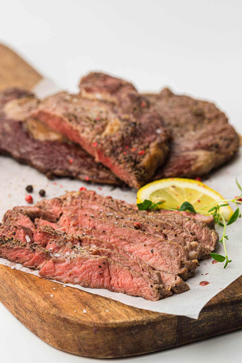 Sliced medium-rare air fryer steak seasoned with pepper on a wooden cutting board with lemon slices and herbs.