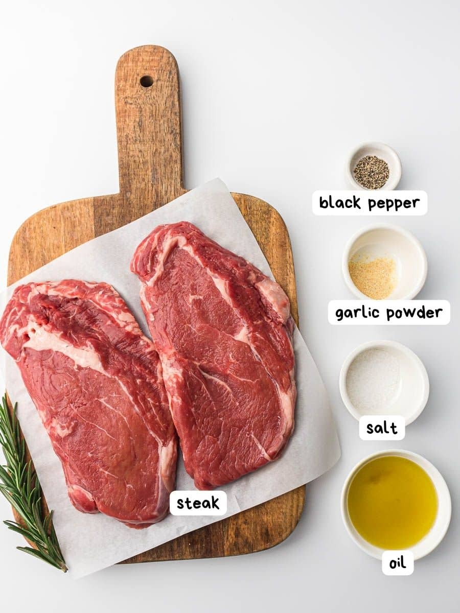Two raw steaks in an air fryer basket with labeled ingredients including black pepper, garlic powder, salt, and oil on a white background.
