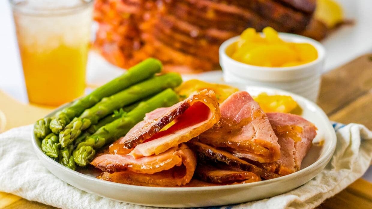 Spiral ham on a plate with asparagus and peaches in the background.