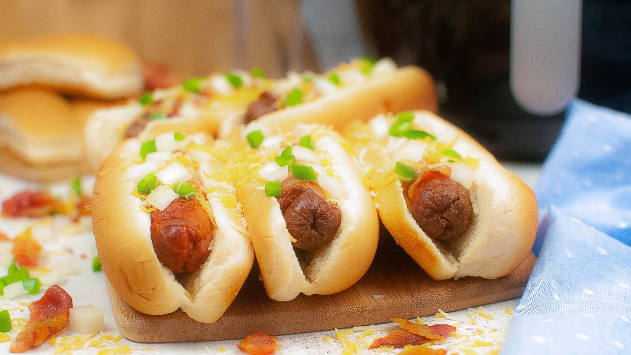 Three bacon wrapped hot dogs in buns topped with cheese and onions.