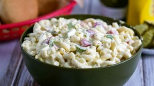 Macaroni salad in a green bowl in front of a hamburger buns and condiments.
