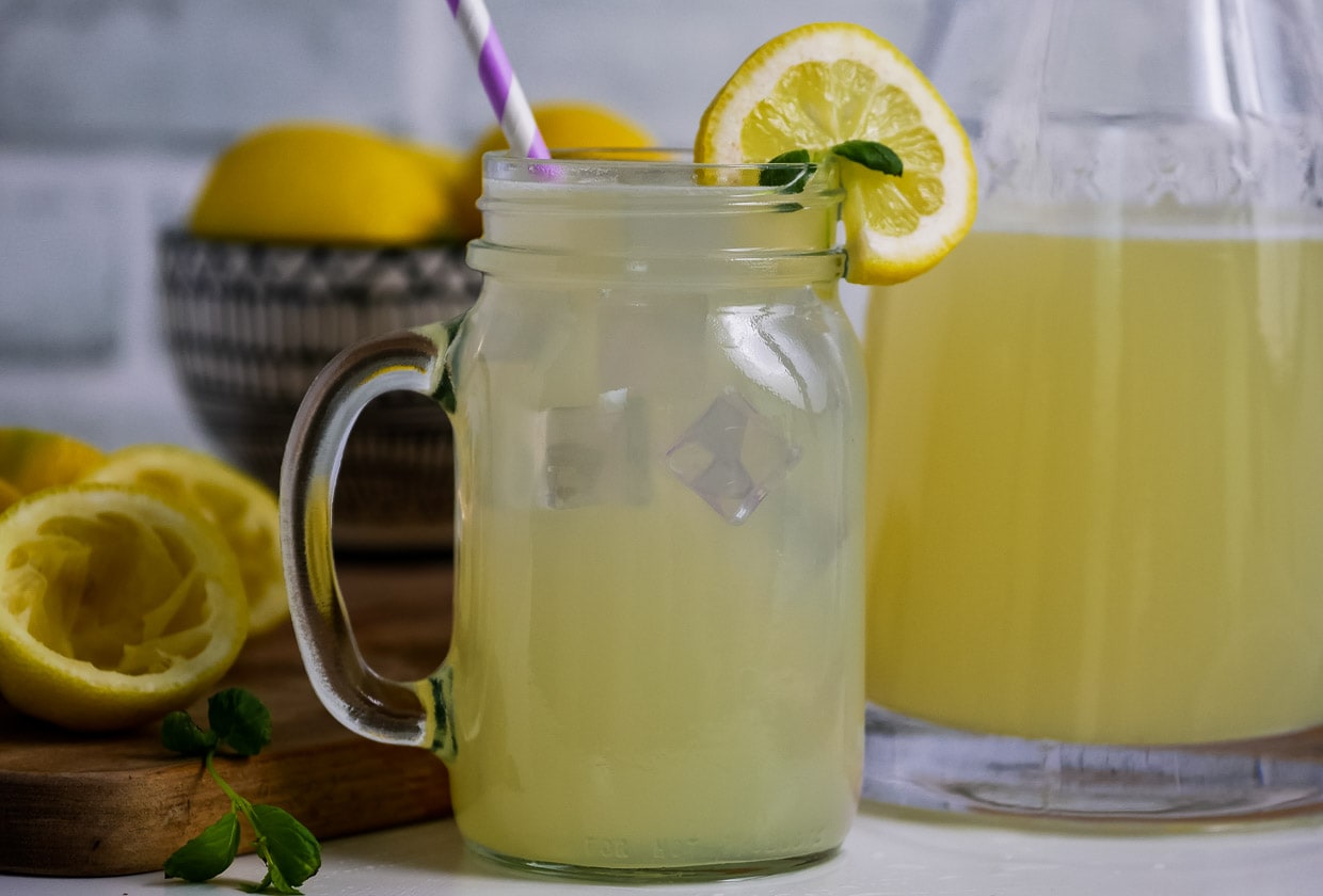 Glass of lemonade on a table with a bowl of lemons in the background and pitcher on the side.