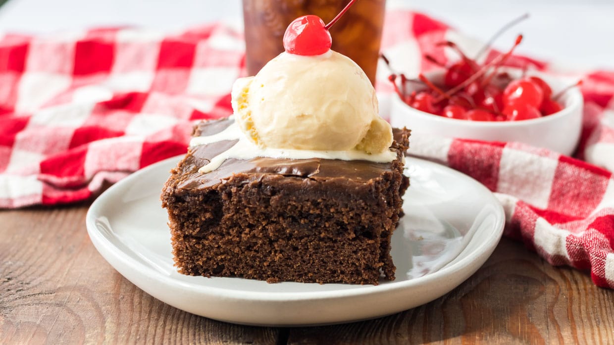 Chocolate coca cola cake on a white plate topped with ice cream and a cherry.