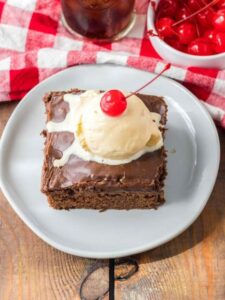 Top view of Coca Cola cake on a plate with ice cream and a cherry and a glass of coke and bowl of cherries in the background.