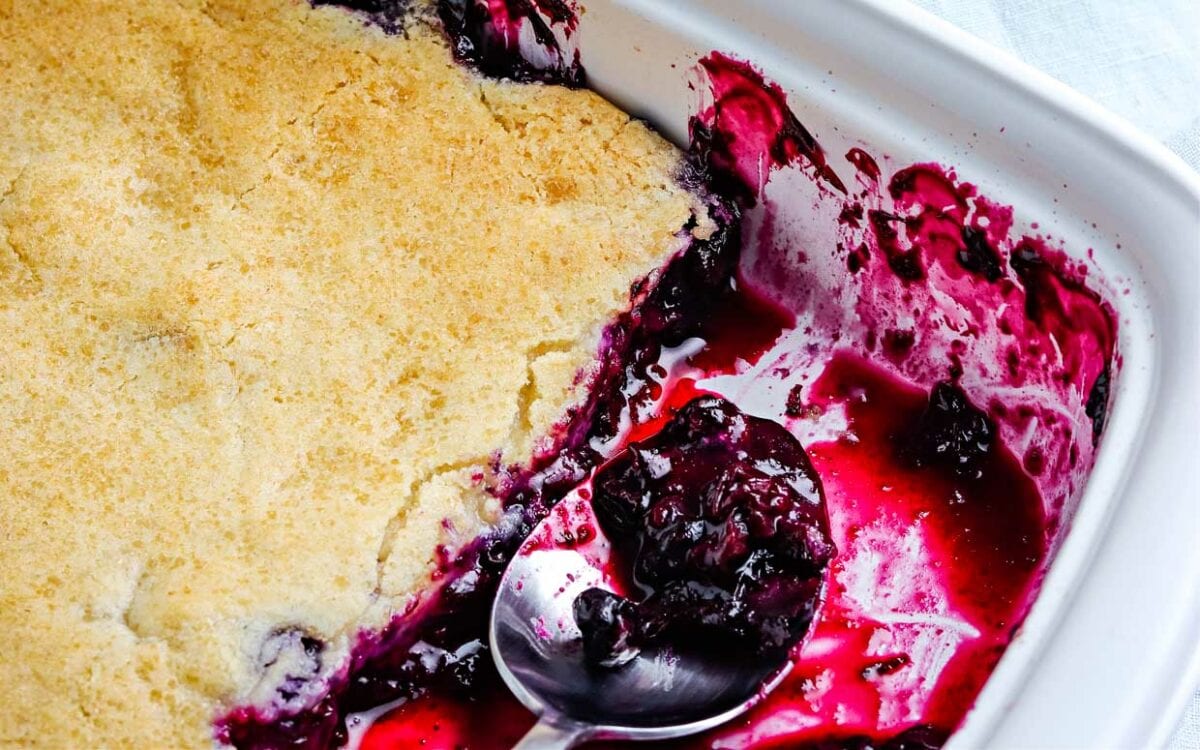 A baking dish with partially served blueberry cobbler, showing a golden biscuit topping and a spoon resting in the purple fruit filling.