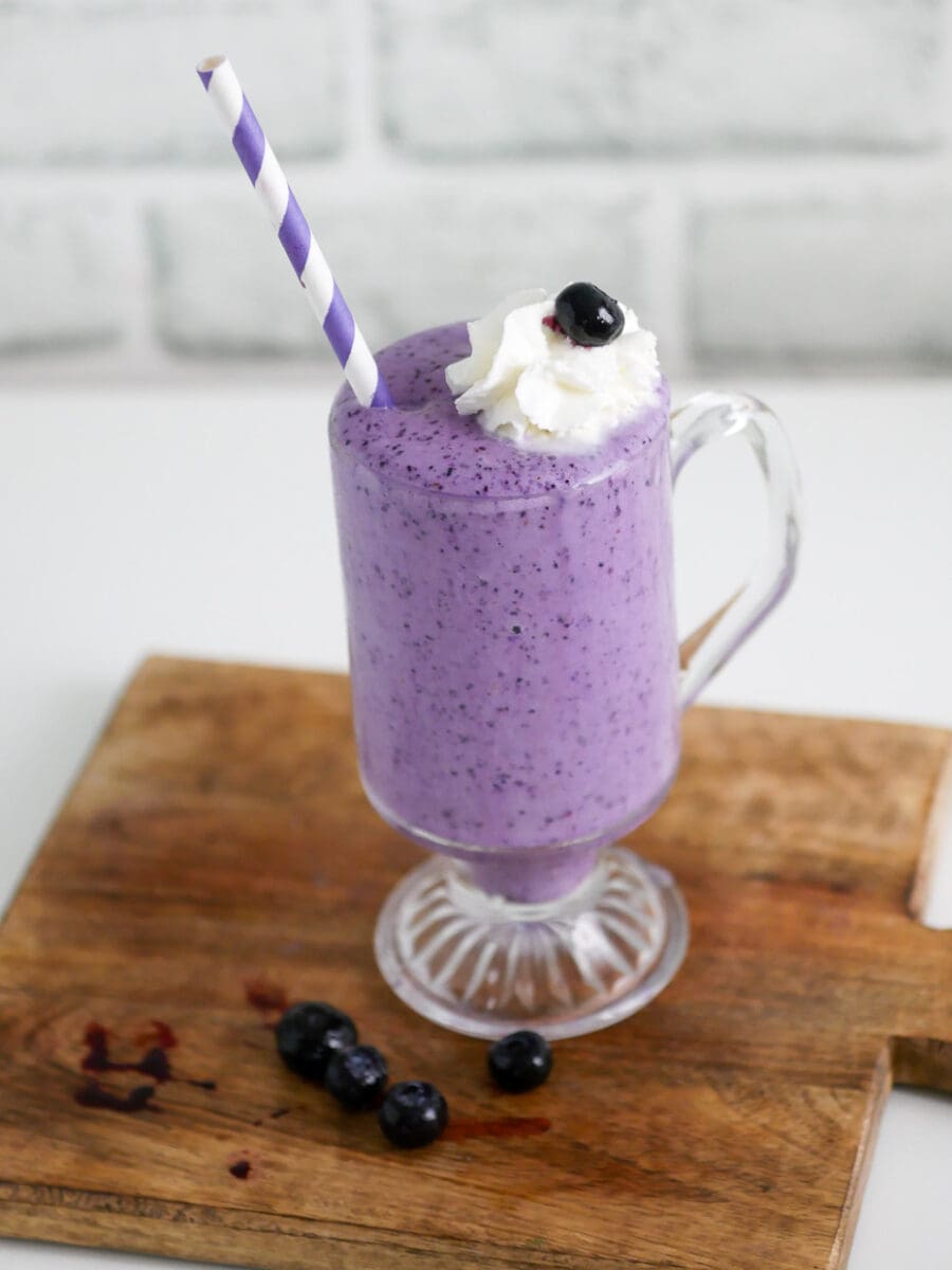 Glass of blueberry milkshake on a wooden cutting board with blueberries.