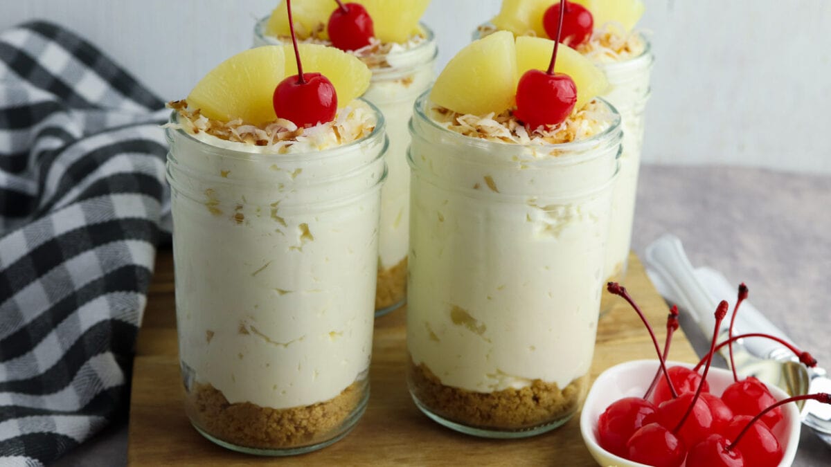 Jars of no bake pina colada cheesecake on a cutting board with cherries, coconut and pineapple.