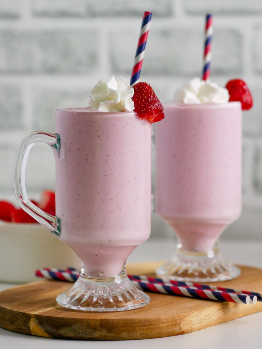 Side view of two glasses of banana strawberry shake on a wooden cutting board with whipped cream and a berry.