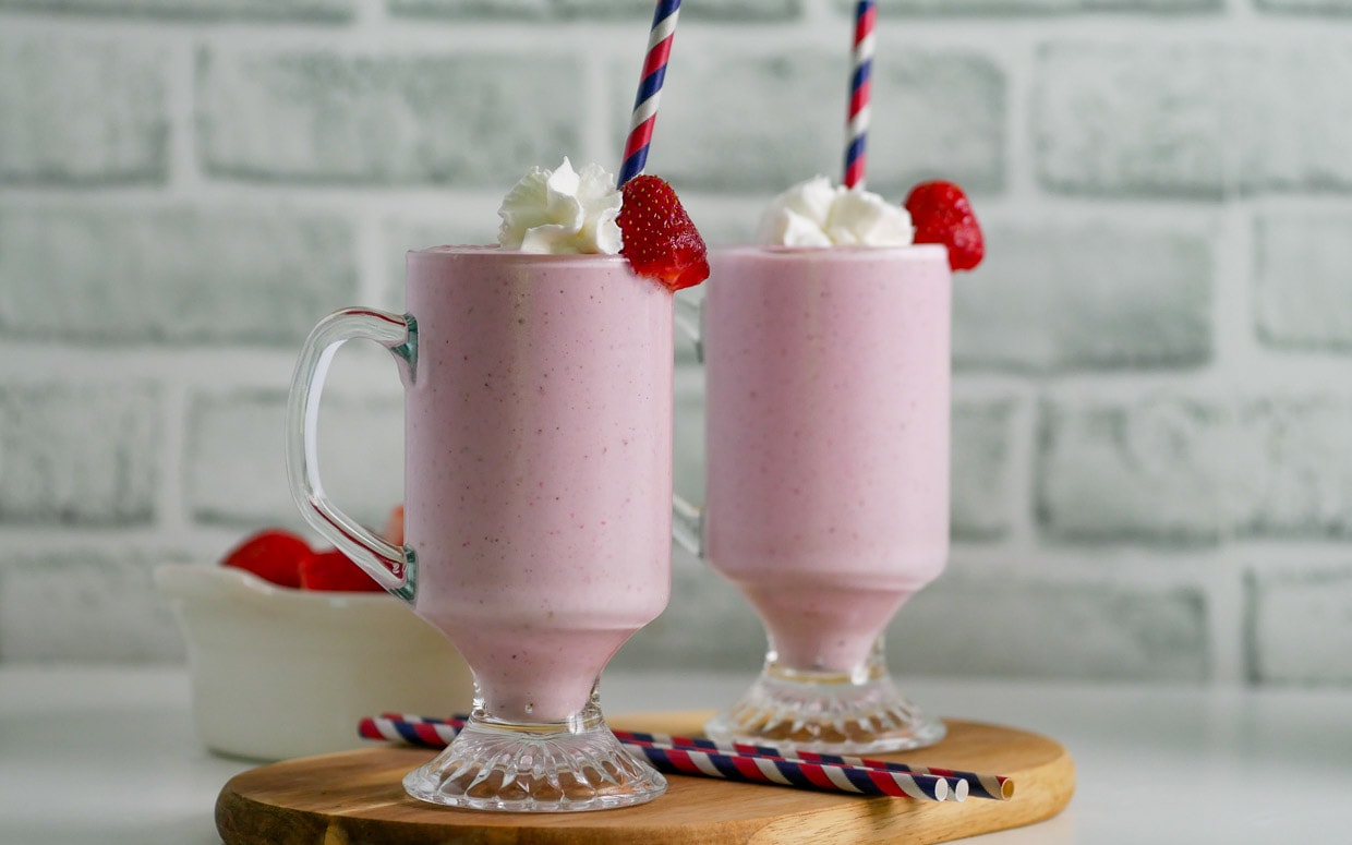 Two glasses of strawberry banana milkshake topped with strawberry and whipped cream on a wooden cutting board with colorful straws.