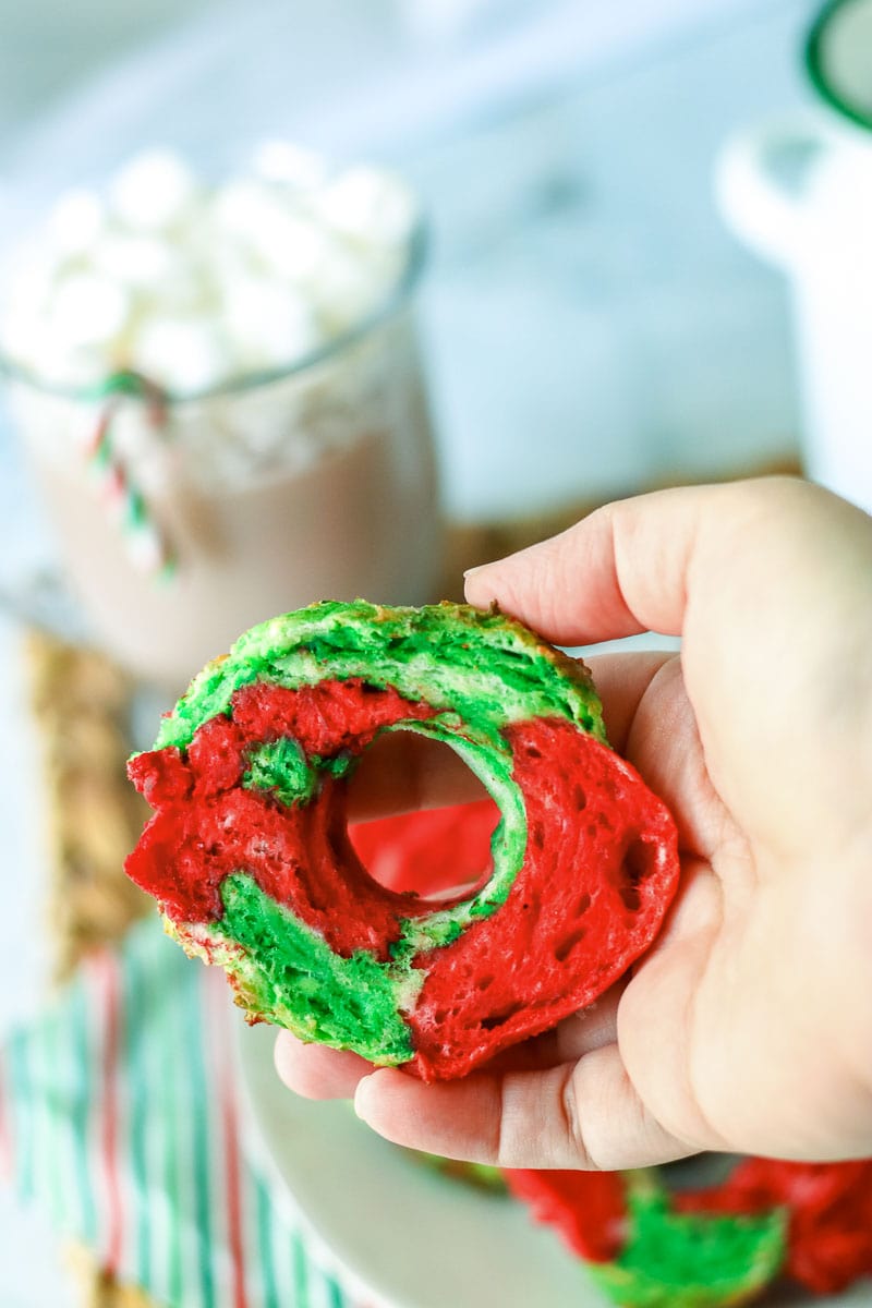 A person holding a doughnut with red and green frosting, resembling Christmas bagels.
