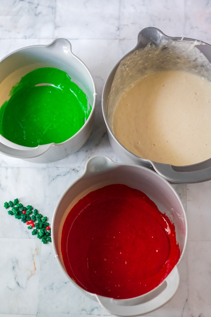 Three bowls of red, green, and white icing on a marble counter, ready to decorate a Christmas bundt cake.