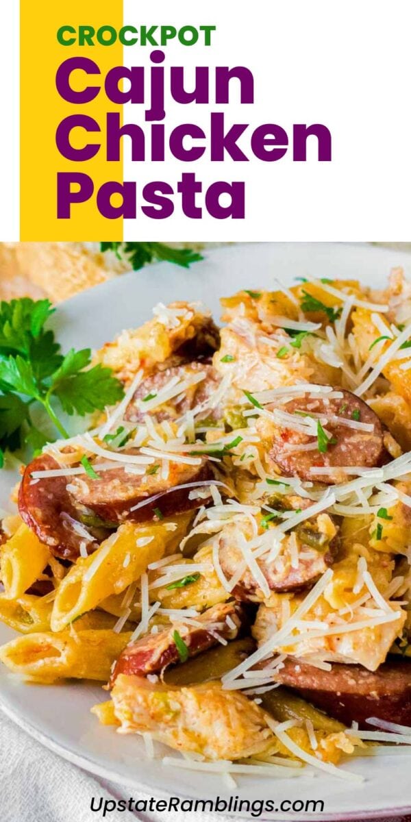 A plate of slow cooker Cajun chicken pasta with sausage, penne, and shredded cheese, garnished with parsley. Text reads “Crockpot Cajun Chicken Pasta UpstateRamblings.com.”.