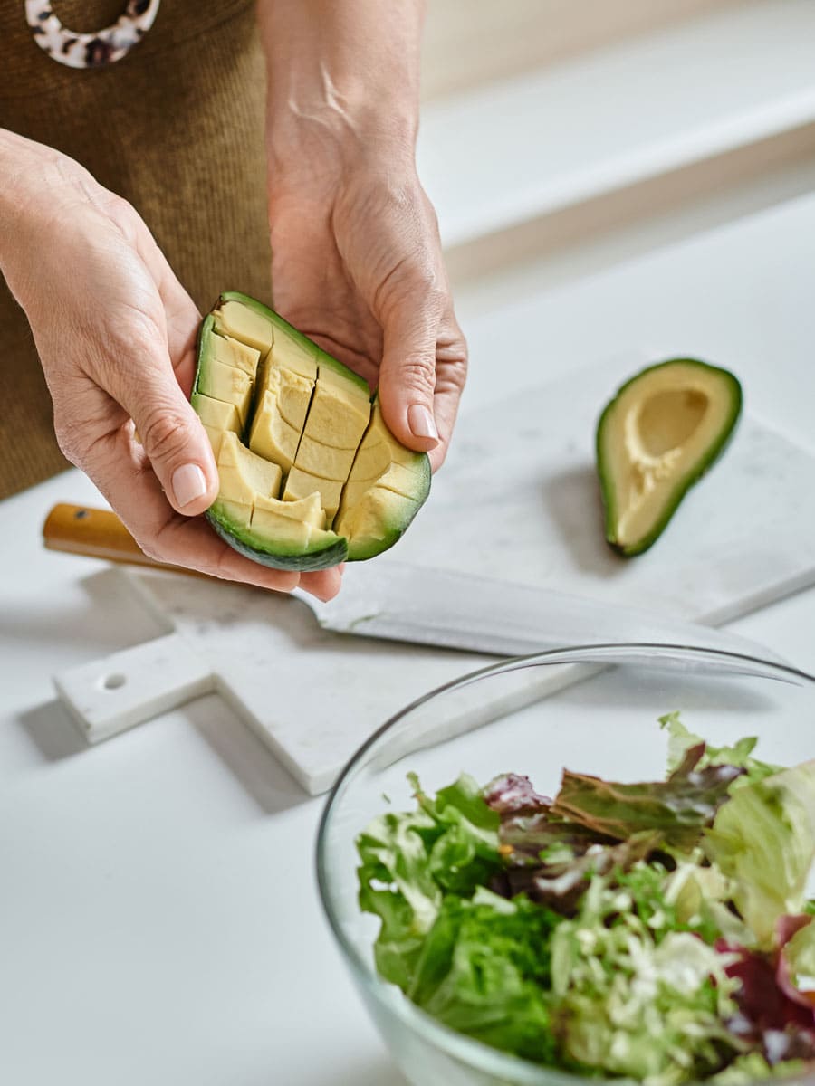 A woman skillfully cuts an avocado into a fresh bowl of salad, demonstrating how to buy and prepare avocados for a healthy meal.