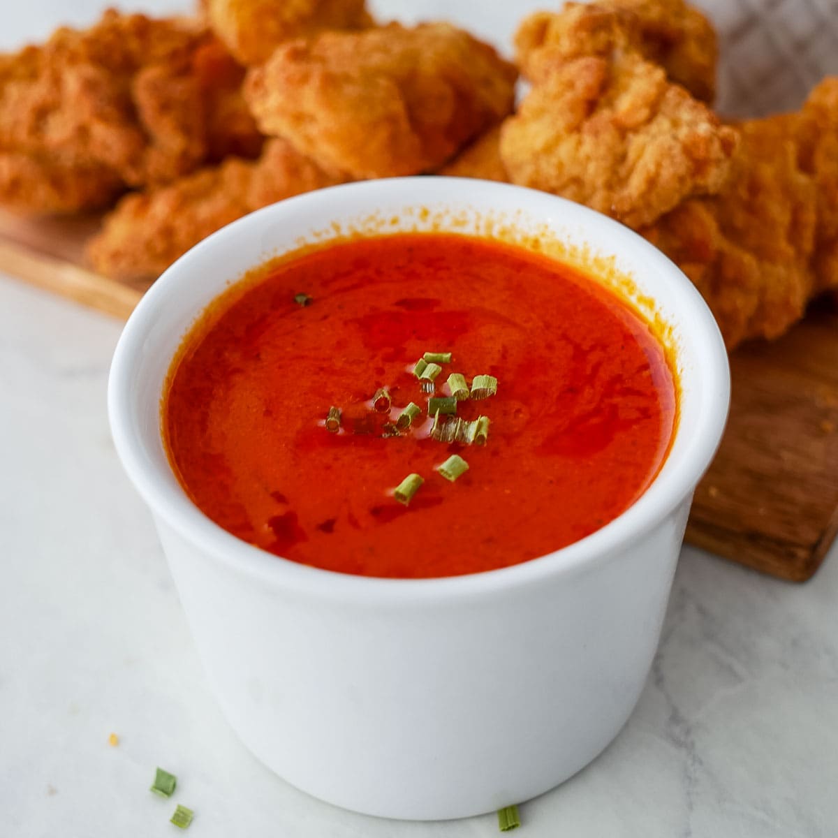 A bowl of buffalo sauce with fried chicken on a cutting board.