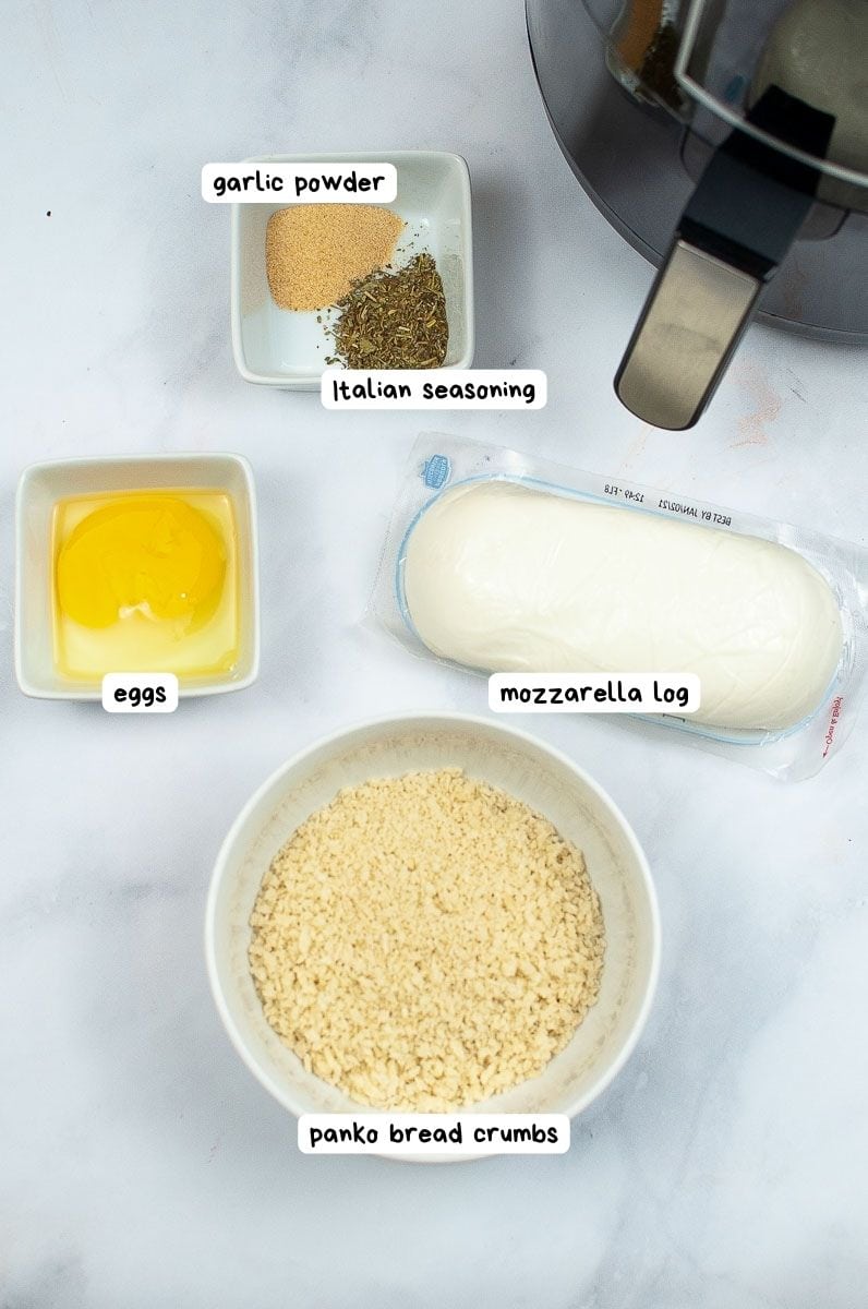 Ingredients for a fried mozzarella laid out on a kitchen surface, including eggs, garlic powder, italian seasoning, mozzarella, and panko bread crumbs.