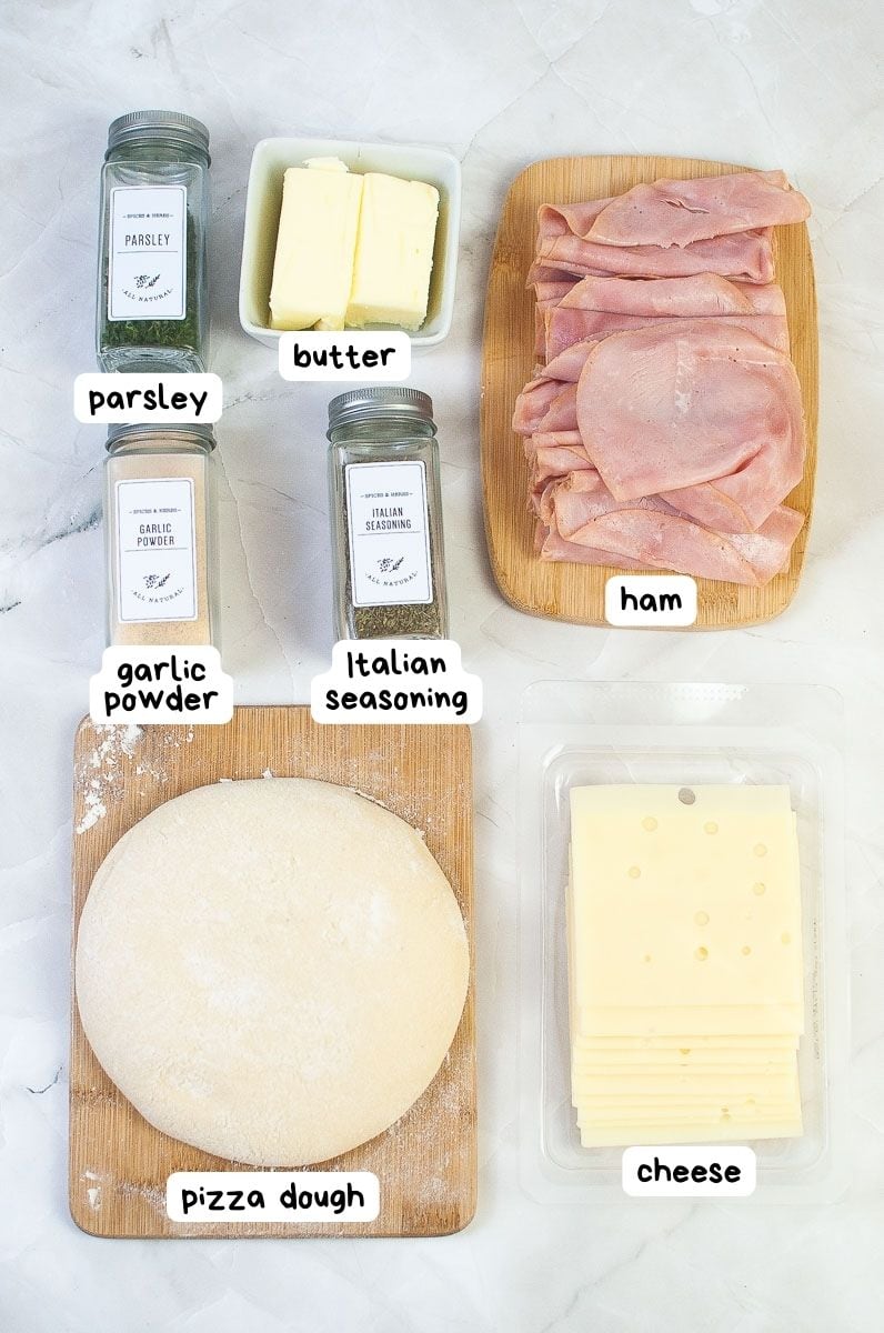 Ingredients for making a ham and cheese pizza rolls laid out on a countertop, including parsley, butter, garlic powder, italian seasoning, ham, pizza dough, and cheese.
