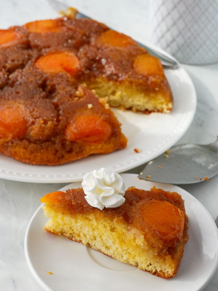 A slice of apricot upside-down cake served on a plate with a dollop of whipped cream, with the rest of the cake in the background.