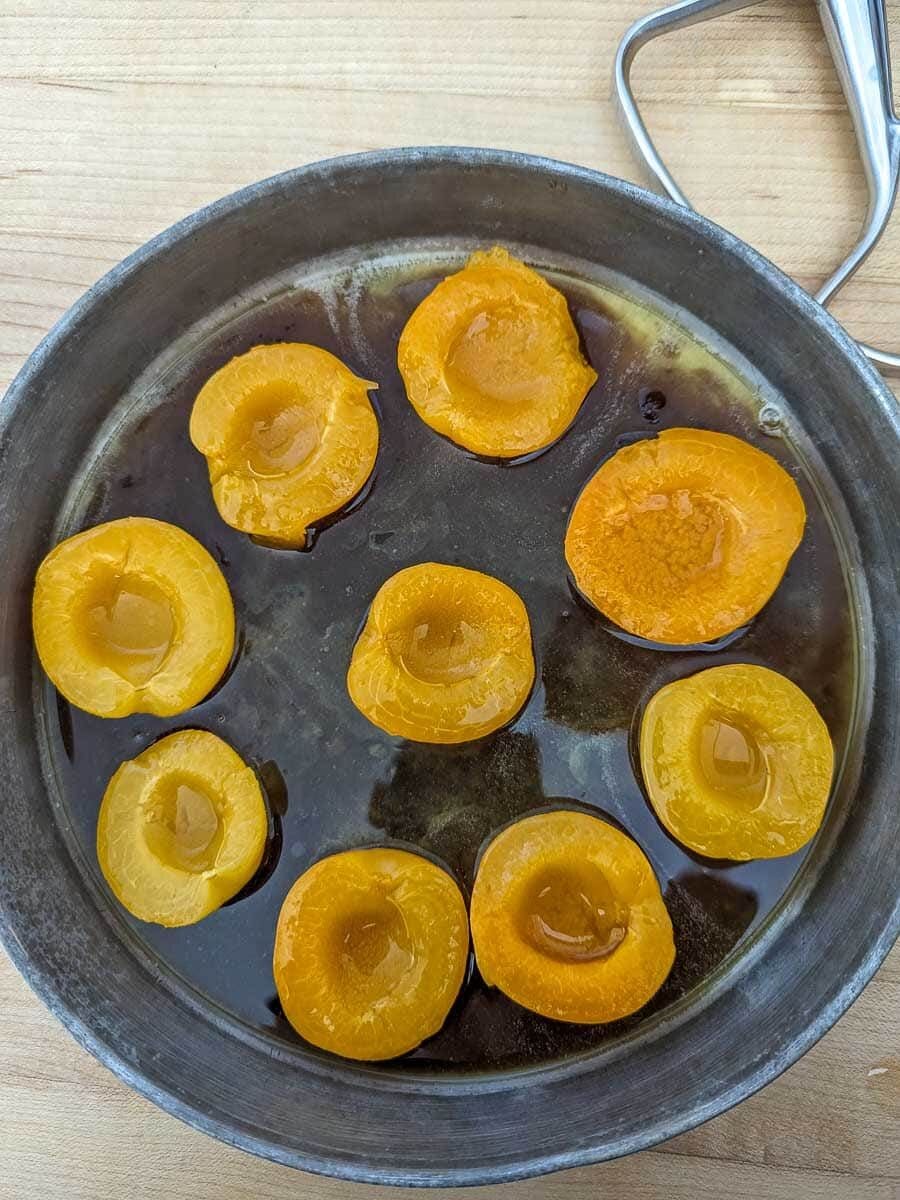 Peeled apricot halves poaching in syrup in a metal pan.