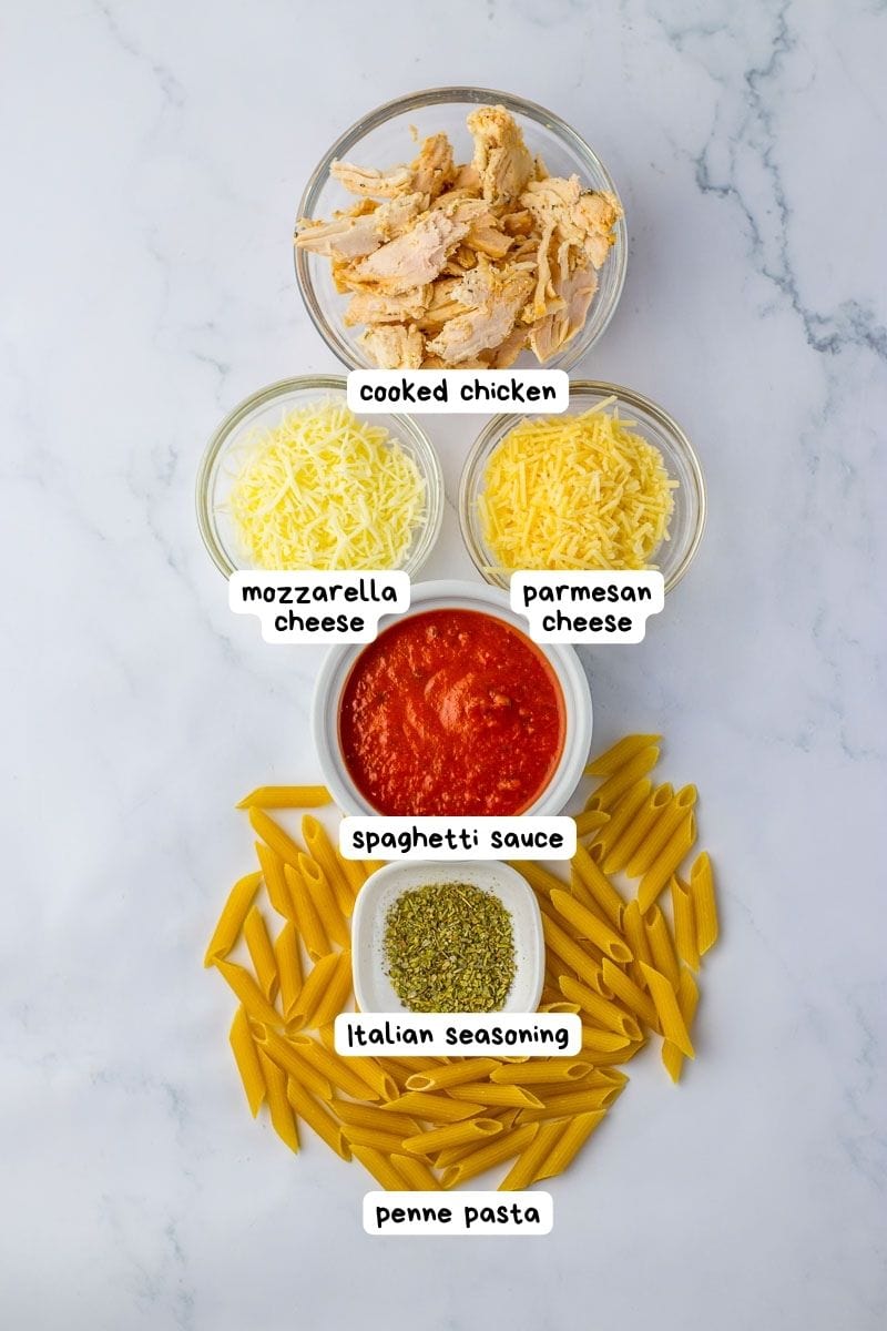 Ingredients for a chicken parm casserole laid out on a marble countertop, labeled with their names