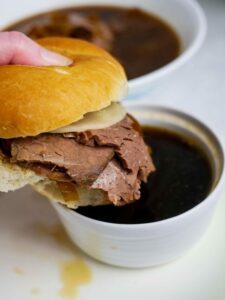A hand holding a beef dip sandwich over a bowl of au jus sauce.