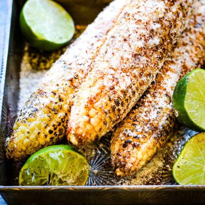 Grilled corn on the cob with seasoning and lime wedges on a tray.